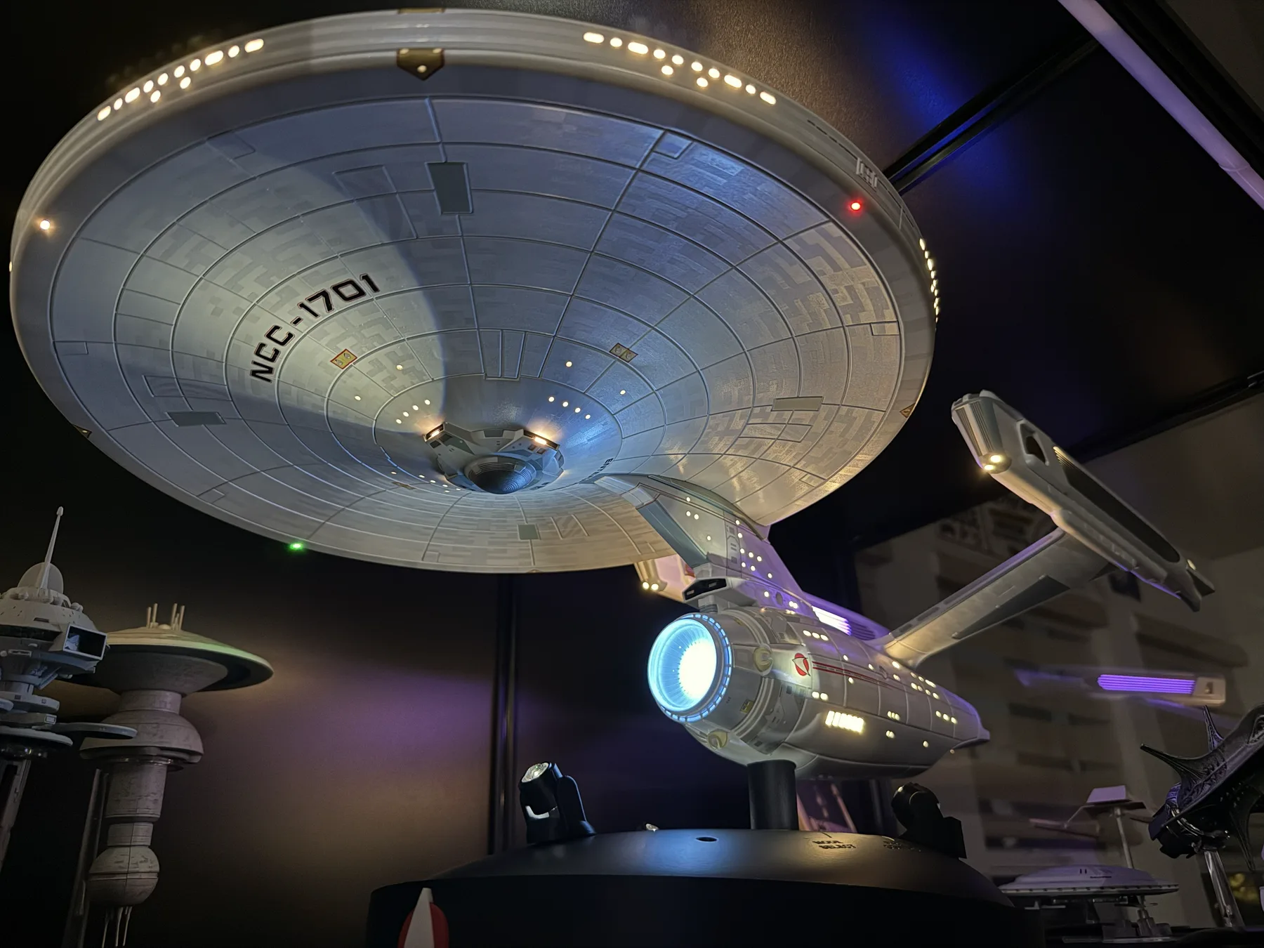 A lit 1/350 starship model displayed on a base, deflector and spotlights glowing in a darkened room.