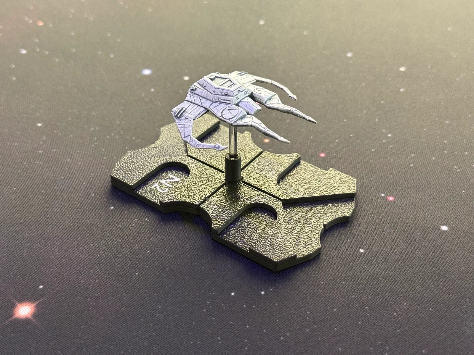 Painted 3D-printed miniature starship, a gray angular hull with long thin spindly legs, on a black printed base with clear peg, on a starfield play mat.